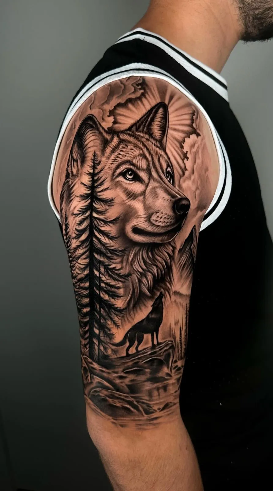 Black and grey realism half sleeve of a wolf portrait against a full moon, a pine tree and a howling wolf below, on the upper arm.